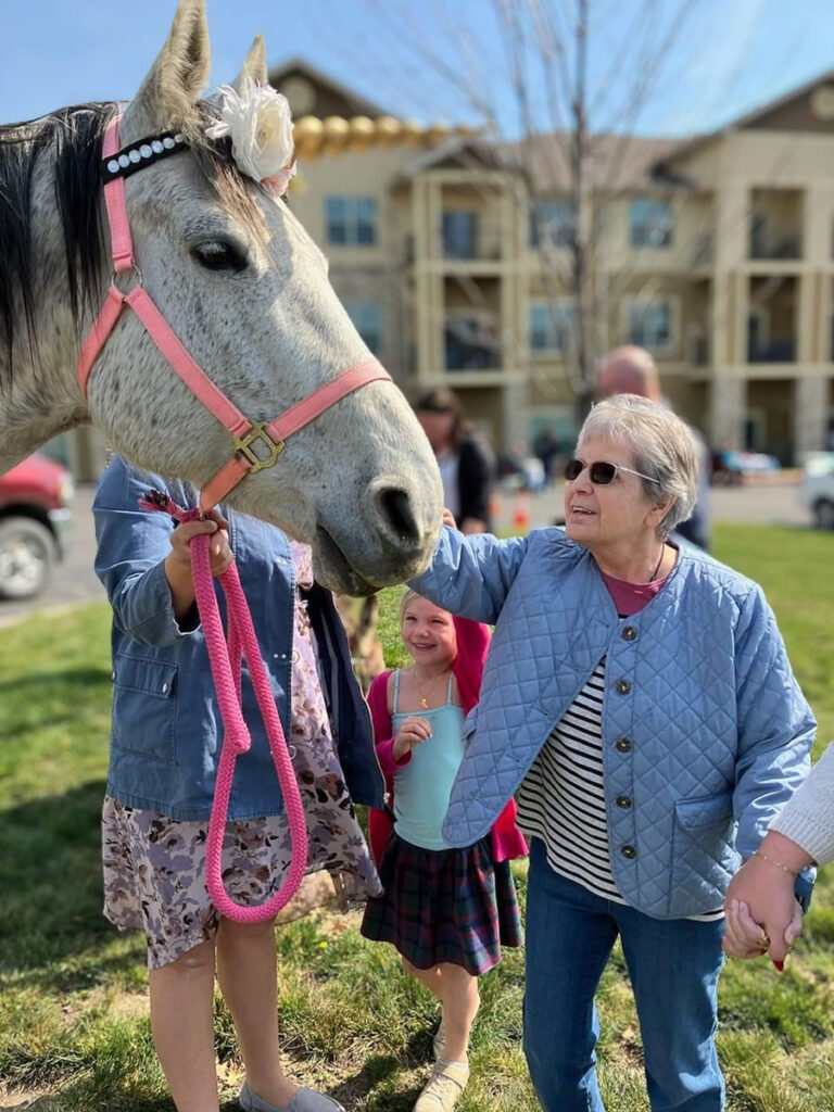 A memory care resident in a blue jacket and sunglasses pets a horse (golden unicorn) at a community event with kids.