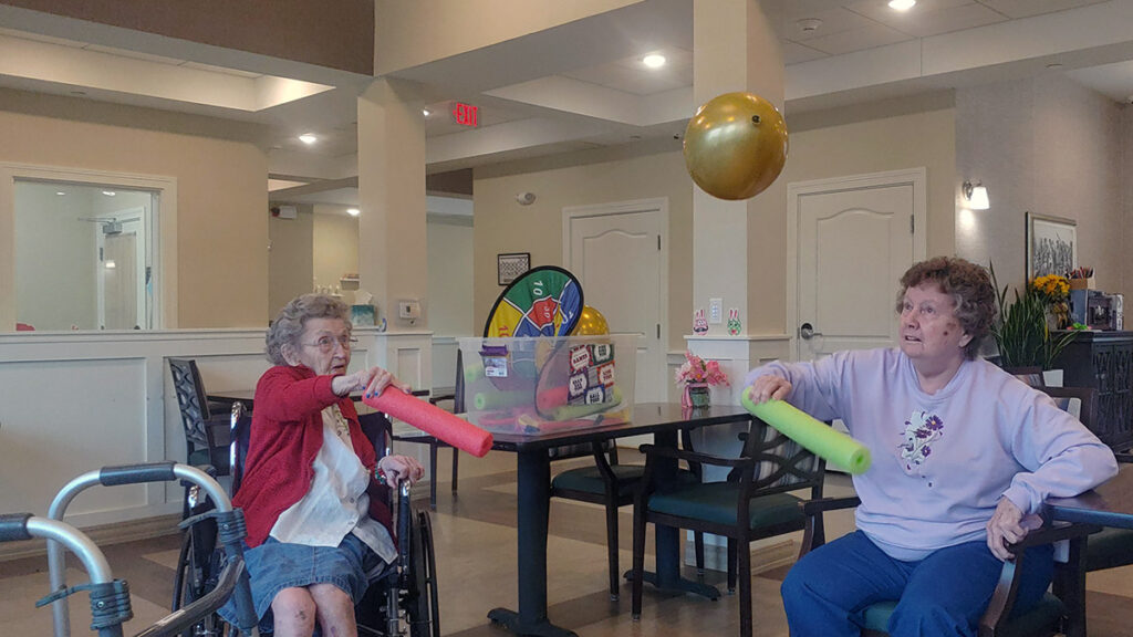 Two happy elderly women are sitting in chairs playing balloon volleyball in the memory care neighborhood.