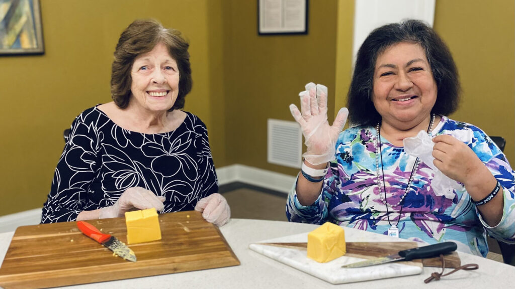 At a table, two women with gloves carefully slice cheese on cutting boards.