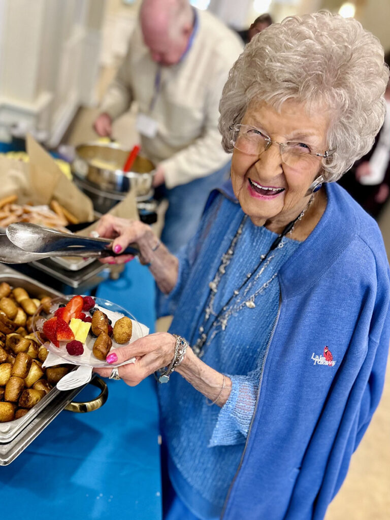 A radiant senior woman in a blue outfit beams with joy at the restaurant buffet in the senior living community. Pure happiness!