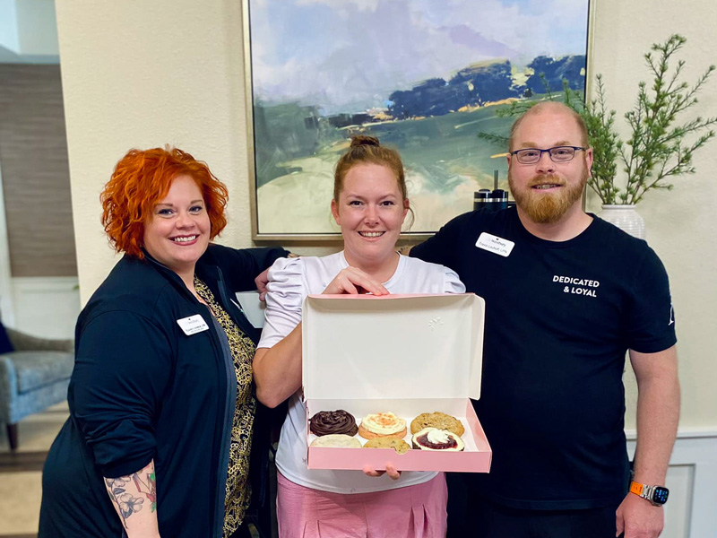 Emily, the Memory Care Director at The Westbury Senior Living, stands with two colleagues, holding a box of Crumble Cookies to celebrate Nurses Appreciation.