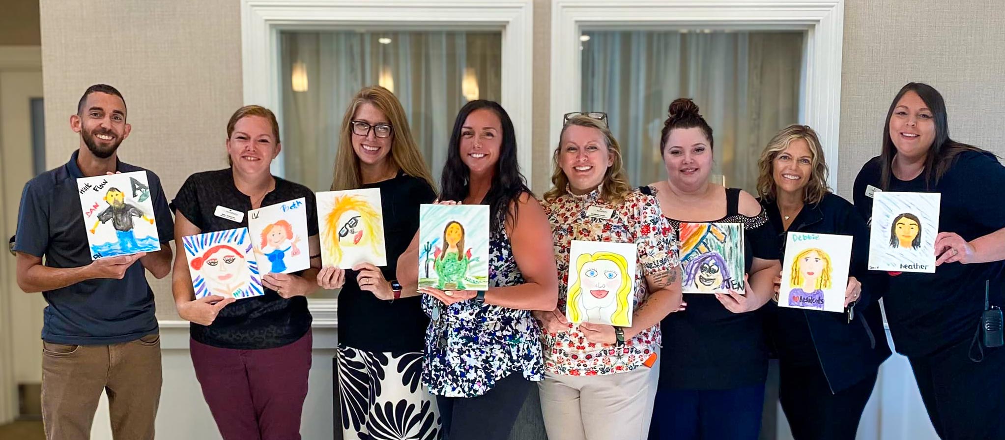 Department heads at The Westbury Senior Living proudly display their self-painted artwork during a team-building activity, smiling and standing in a line.