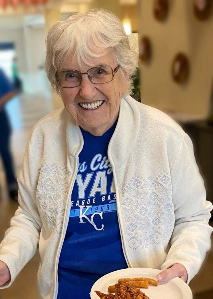 Smiling senior resident wearing a Kansas City Royals shirt holds a plate of food. She is dressed in a light jacket, standing in a well-lit senior living community.