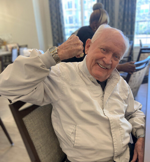 Smiling senior resident in a white jacket raises his arm in a playful flexing gesture while seated in a senior living dining area, exuding confidence and joy.