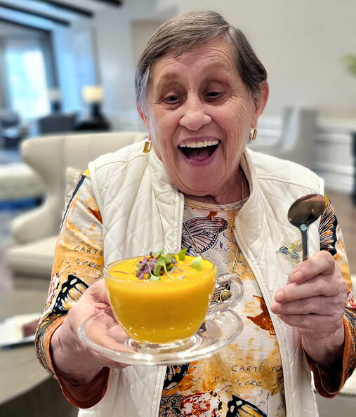 Smiling senior resident joyfully holds a bowl of vibrant orange soup garnished with greens, wearing a colorful butterfly-patterned shirt and white vest, ready to enjoy the meal.