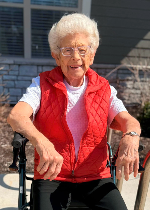 Smiling senior resident wearing a red quilted vest enjoys the sunshine while seated outdoors with a walker.