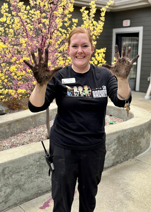 Team member shows off her muddy hands with a big smile after gardening, standing in front of a blooming tree in the courtyard.