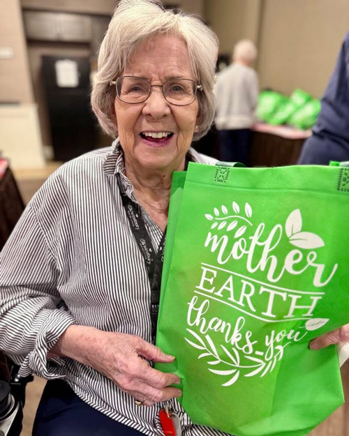 Smiling senior resident holding a bright green reusable tote bag with the words "Mother Earth Thanks You" during an Earth Day celebration.