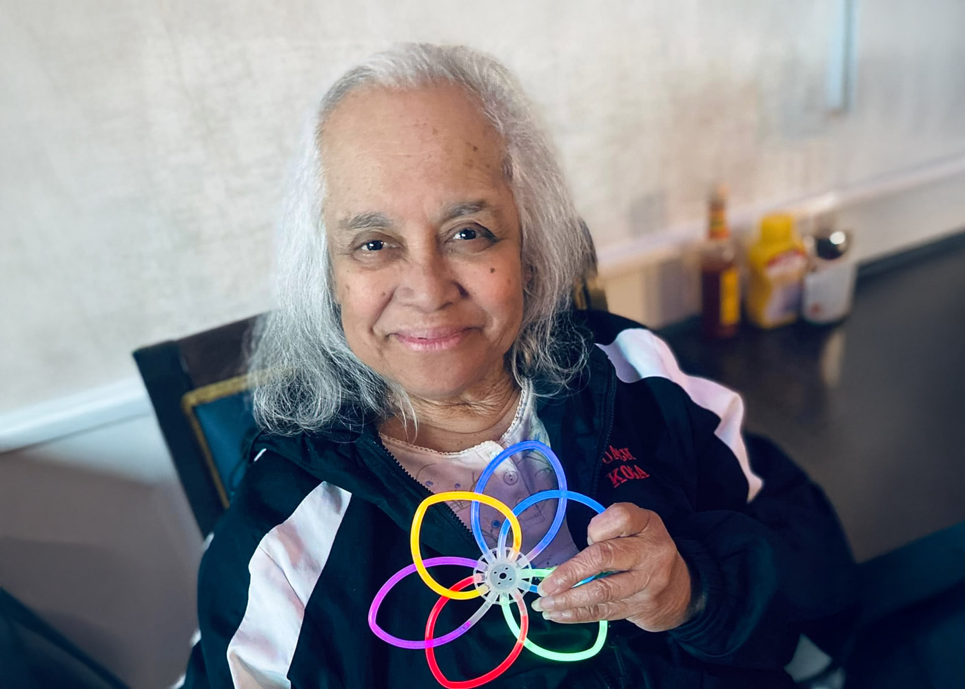 Smiling senior woman holding a colorful interlocking ring flower toy, seated indoors with condiment bottles in the background