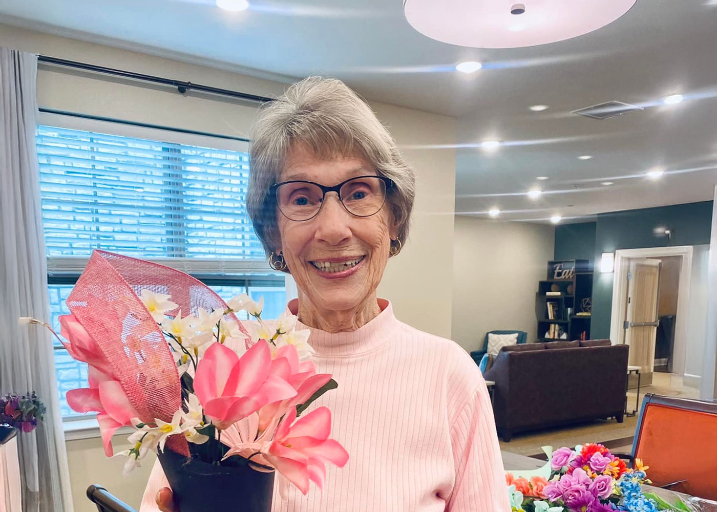 Smiling senior woman in a light pink sweater holding a vibrant arrangement of pink and white artificial flowers inside a well-lit room