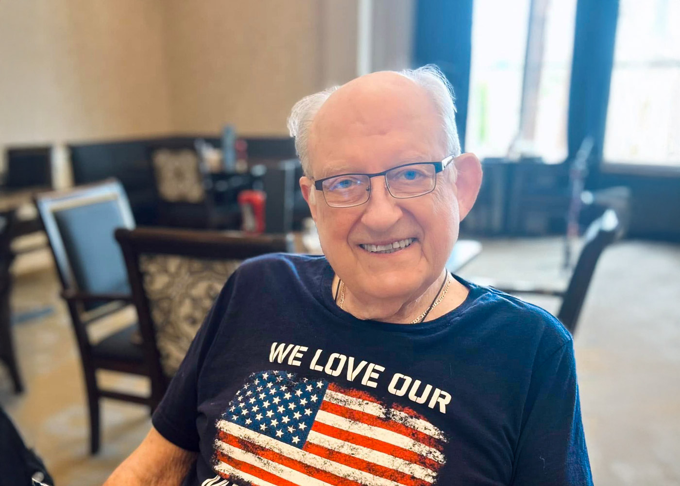 Smiling senior man wearing an American flag heart shirt with the words “We Love Our” while seated in a warmly lit dining room