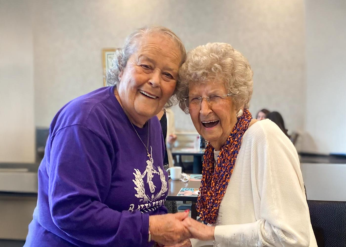 Two joyful senior women smiling and holding hands, one in a purple shirt and the other in a white sweater with an orange and purple scarf