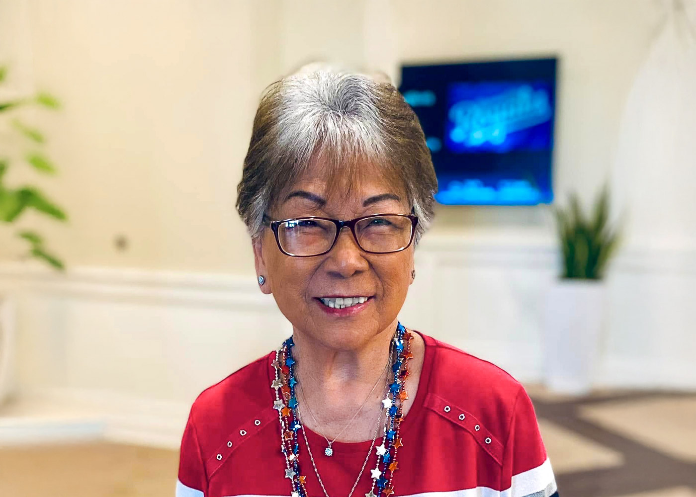 Smiling senior woman wearing red, white, and blue attire with star necklaces, standing indoors with a TV and plants in the background