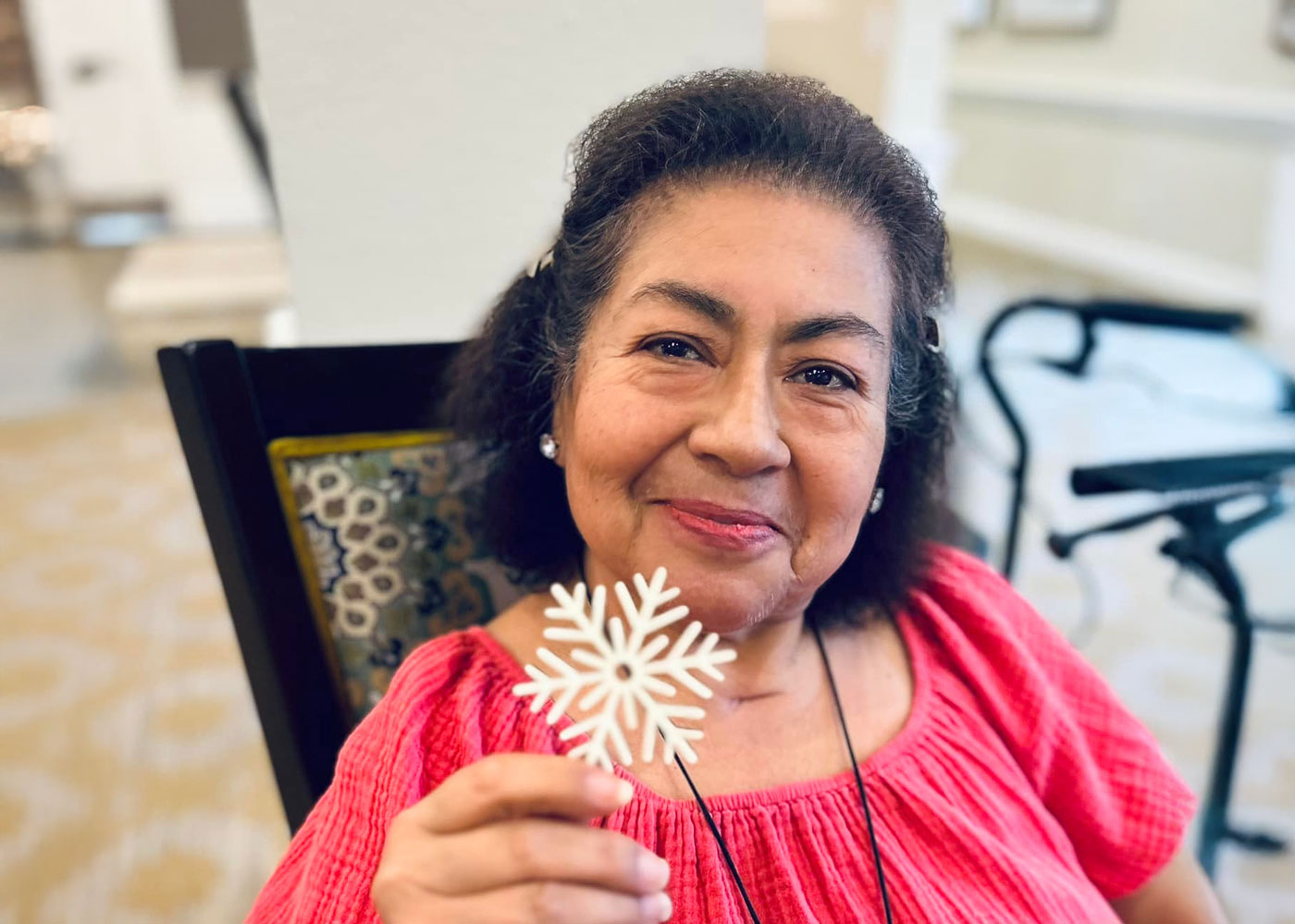 Smiling senior woman in a bright pink top holding a decorative white snowflake, seated indoors near a walker