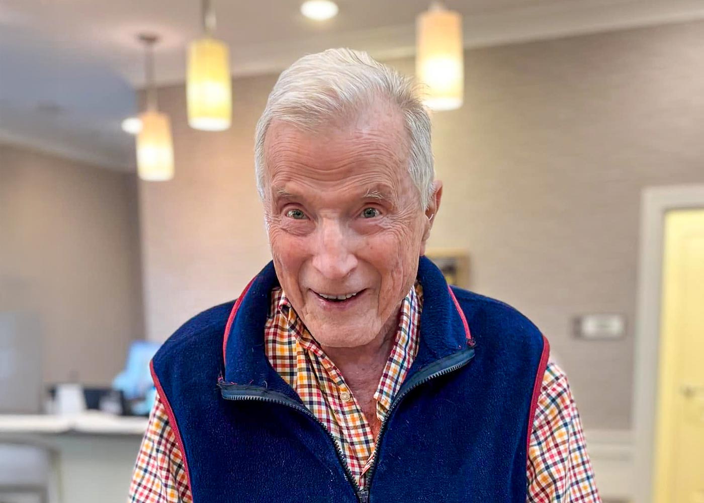 Smiling senior man in a plaid shirt and navy fleece vest standing indoors under warm ceiling lights