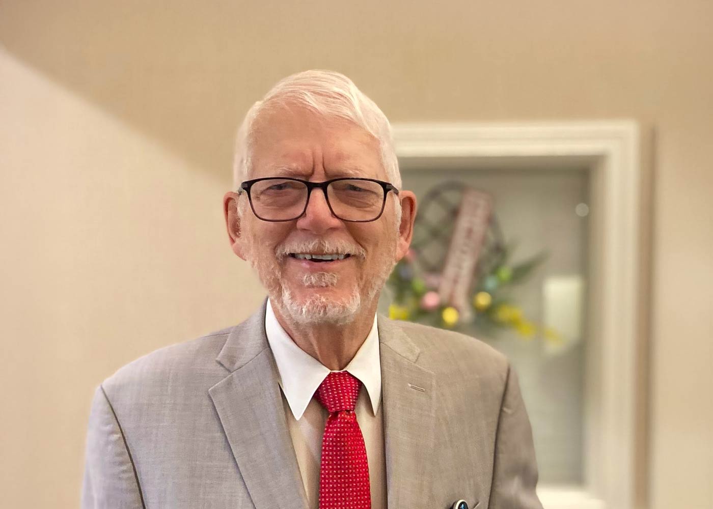 Smiling senior man in a light gray suit and red tie, wearing glasses, standing indoors with a spring floral wreath in the background