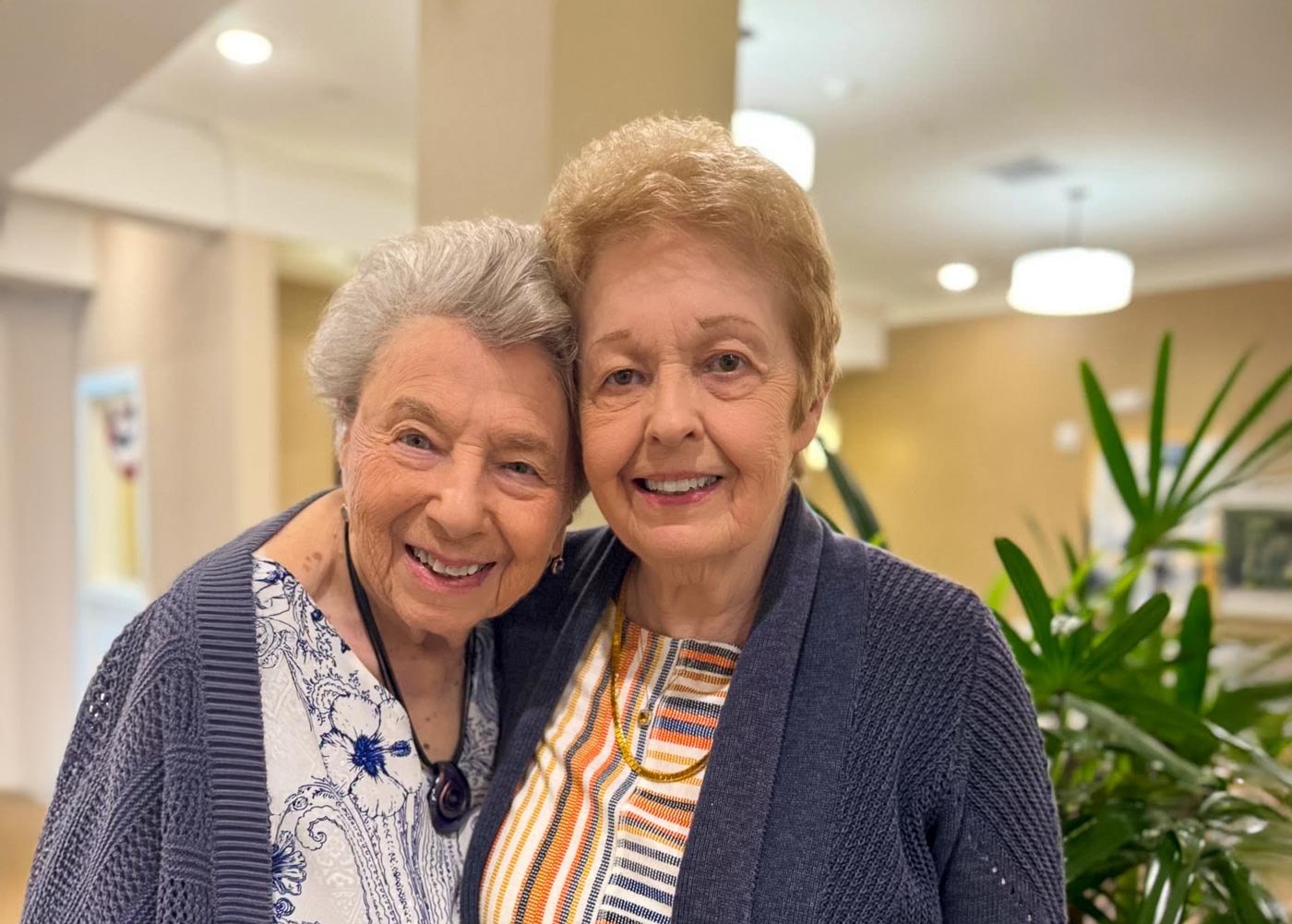 Two senior women smiling and embracing each other inside a warmly lit community space