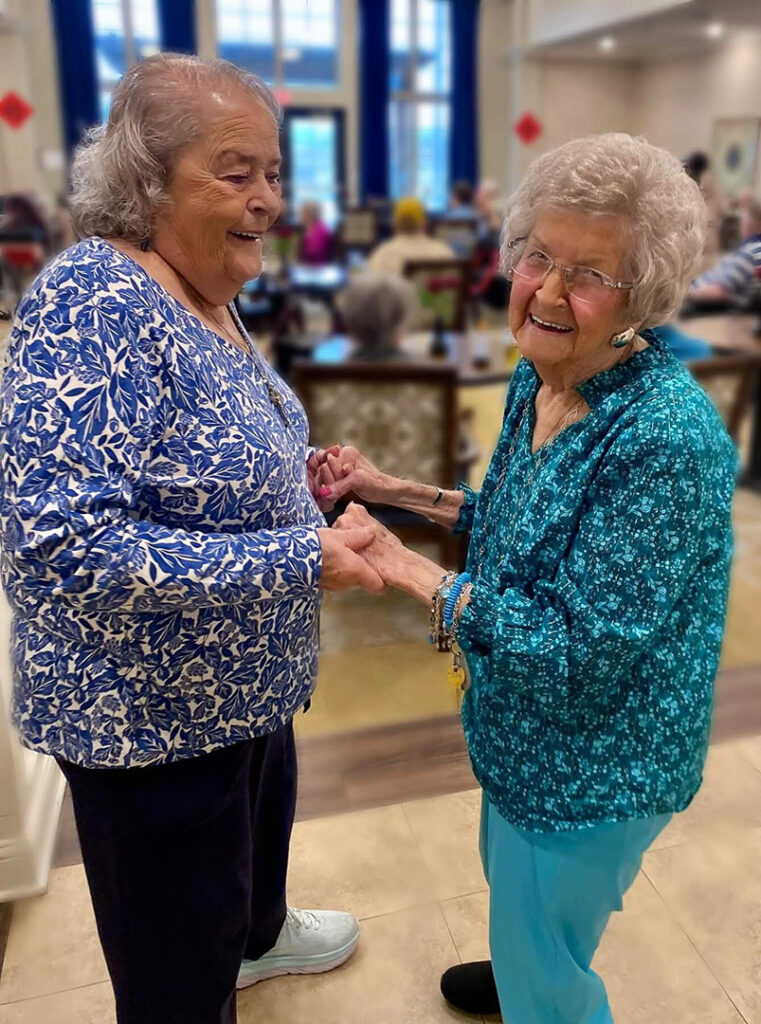 Two smiling senior women dance hand in hand at a community event.