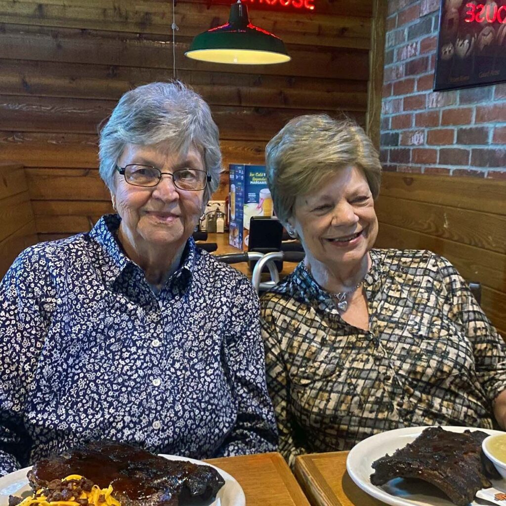 Two senior women sit together at a restaurant booth, smiling with plates of barbecue.