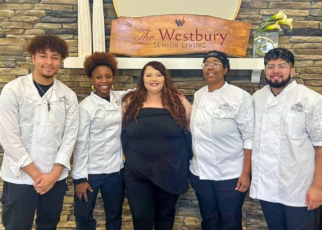 Culinary team at Westbury Senior Living poses together in white chef coats, standing proudly in front of the community sign, showcasing teamwork and hospitality.