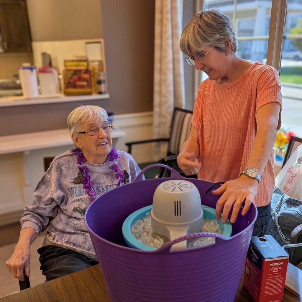 Two women enjoy making homemade ice cream together at a senior living community, smiling as they prepare ingredients in a purple ice cream maker bucket.
