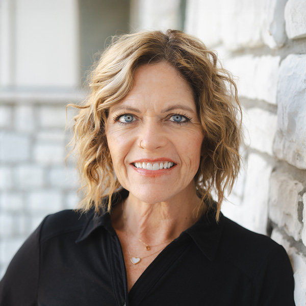 Deborah “Debbie” Hook, Senior Living Director at The Westbury Senior Living, smiling outdoors in a black top with light stone walls softly blurred behind her.