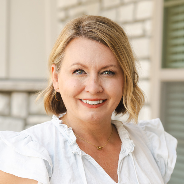 KayMaree Hader, Assistant Executive Director at The Westbury Senior Living, smiling outdoors in a white ruffled top with soft stone and siding in the background.