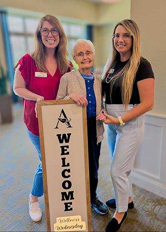 Two team members smile alongside a senior woman, posing with a Welcome sign for Wellness Wednesday.