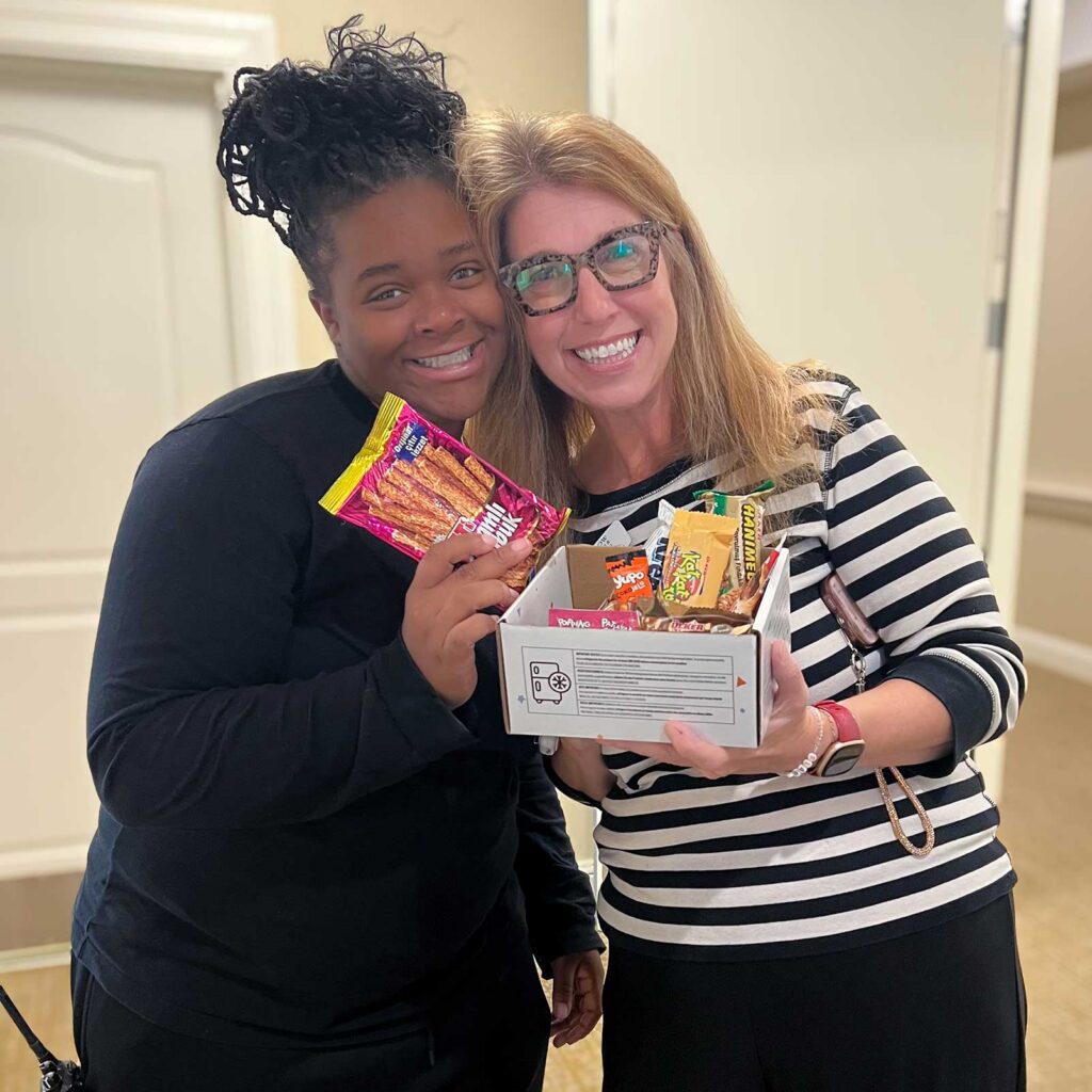 Two team members smile, heads together, holding up a box of treats given in gratitude.