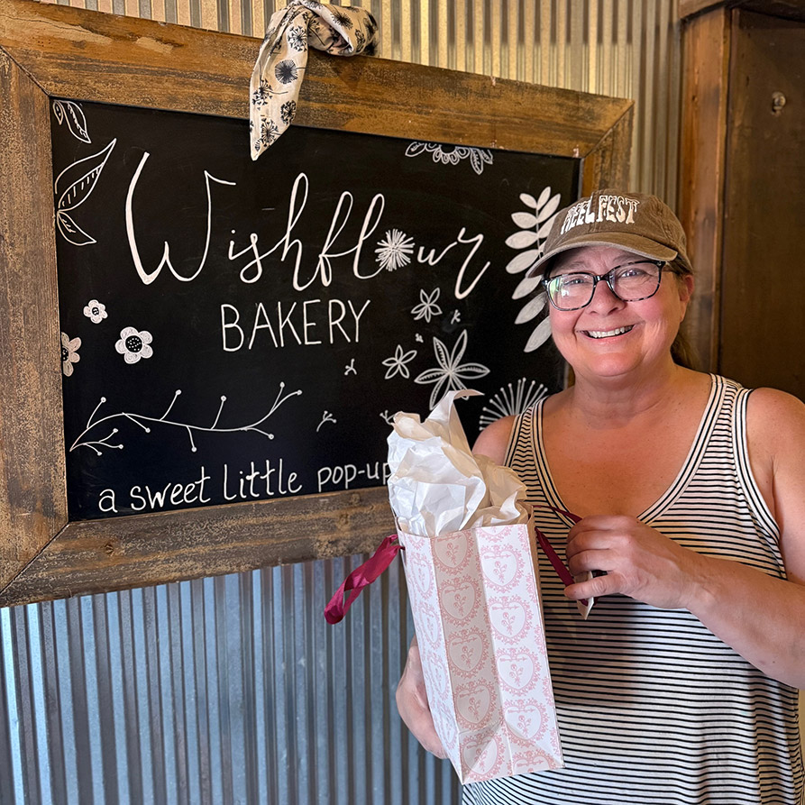A woman smiles beside the sign for Wishflour Bakery with a gift bag in her hand.