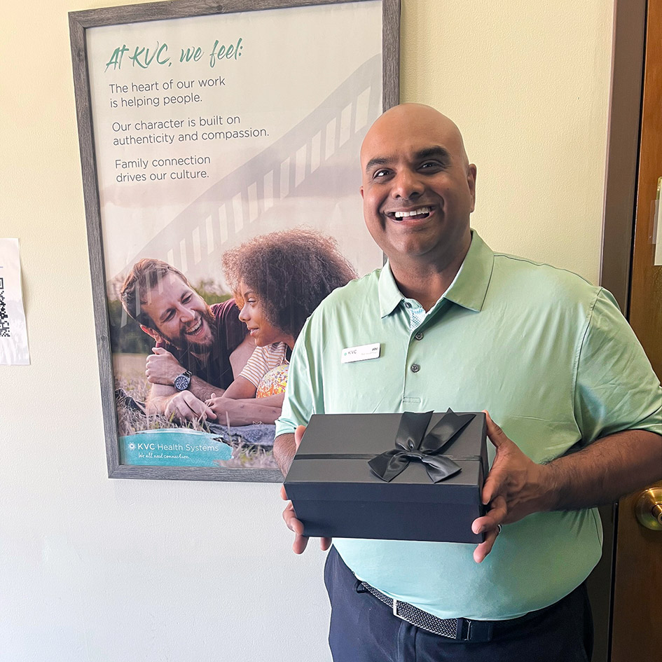A man smiles, holding an elegantly packaged gift, beside a poster for KVC Health Systems.