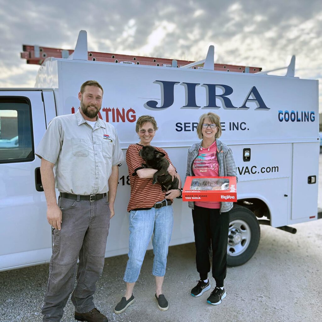 Owners of Jira Heating and Cooling, along with a small dog, smile next to the company work truck, one woman holding a box of doughnuts.