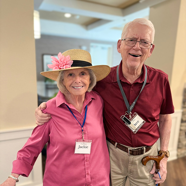 A senior man wearing a maroon shirt and a senior woman wearing a pink blouse and a hat with a large pink flower smile brightly together at The Westbury Senior Living.